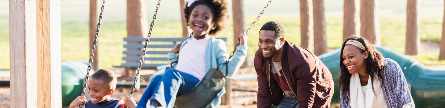 Young Family Playing On Swing Set In Playground