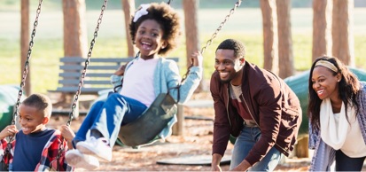 Young Family Playing On Swing Set In Playground